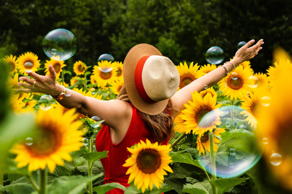 Ženska s slamnikom stoji v polju sončnic z odprtimi rokami